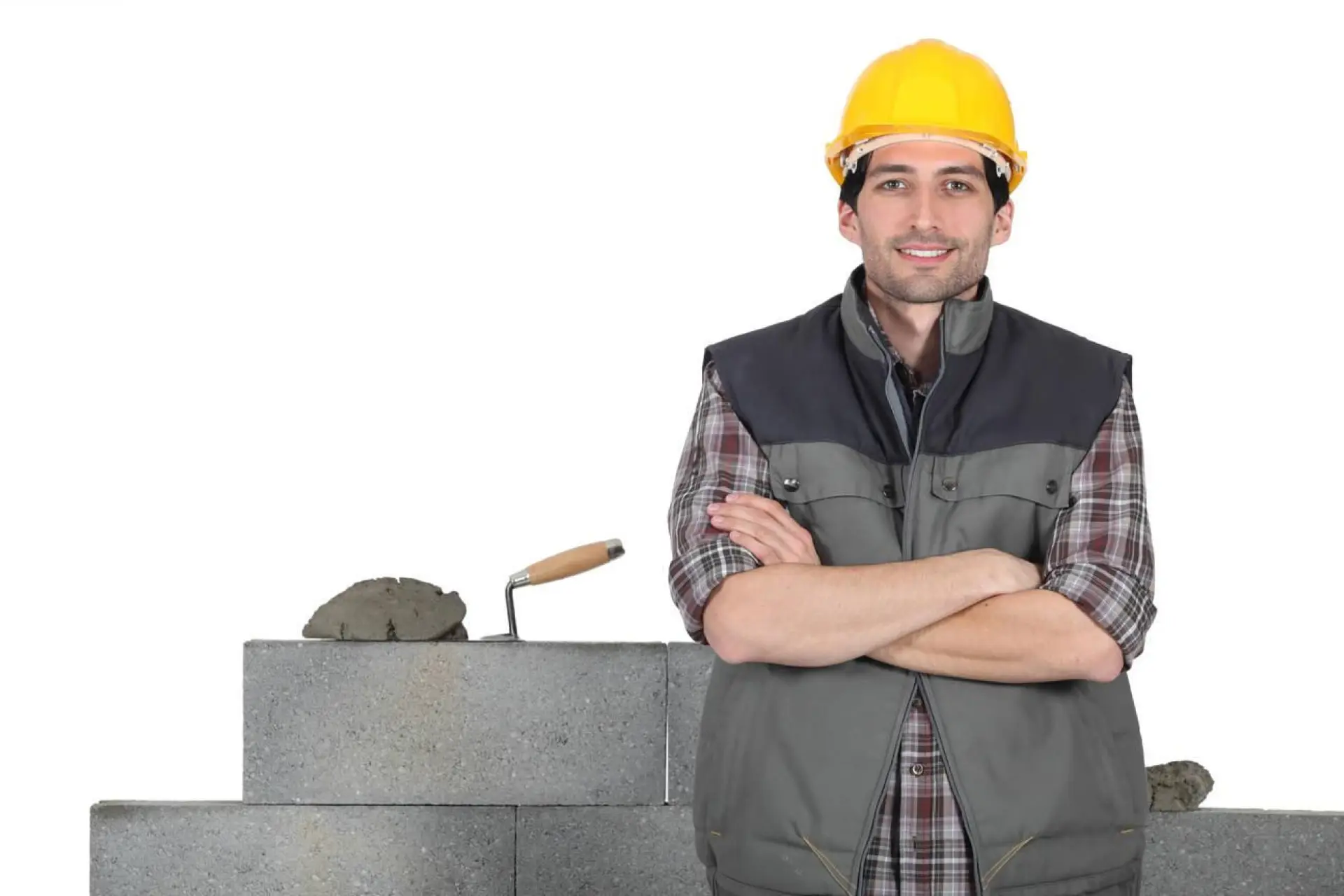 Artisan maçon souriant avec casque de sécurité jaune devant un mur de parpaings avec truelle et mortier
