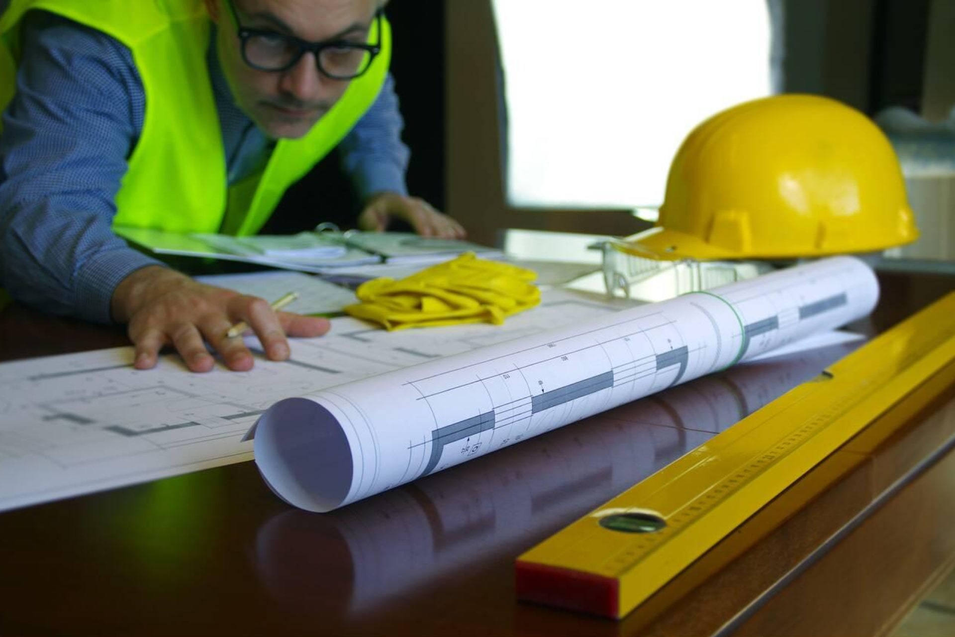 Architecte en gilet de sécurité analysant des plans de construction avec un casque jaune et des gants sur la table.