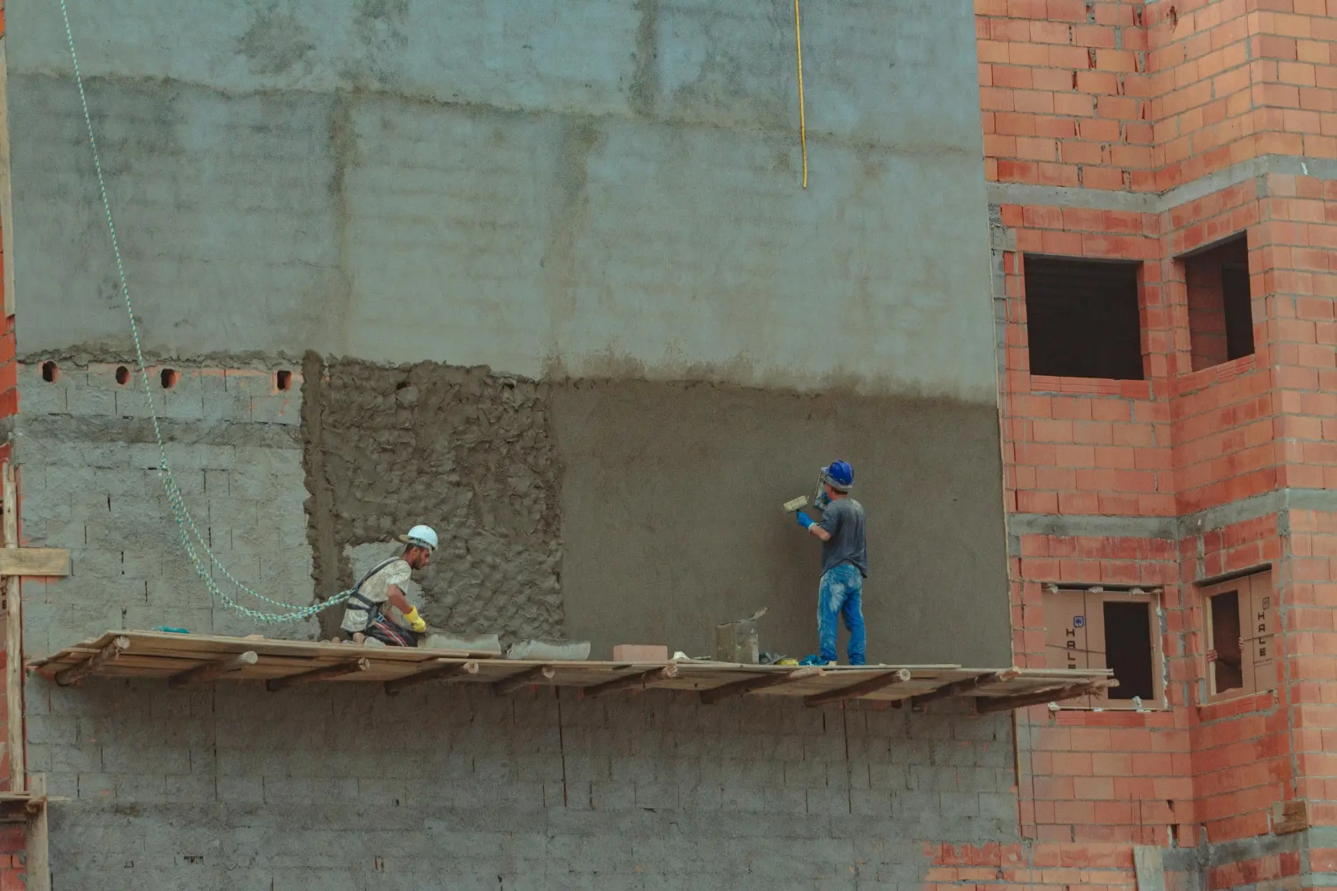 Deux maçons en action, appliquant du plâtre sur le mur extérieur d'une structure en briques, démontrant des techniques de rénovation immobilière.